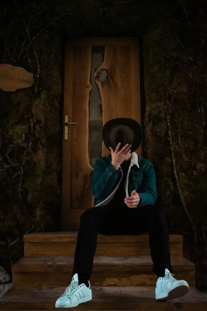 Man in a fashionable outfit sits on steps with a rustic door backdrop, holding a hat