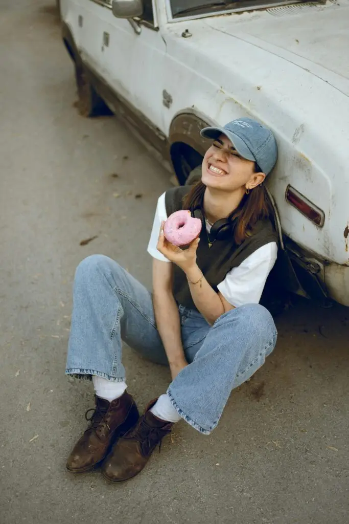 Young woman in casual outfit smiling while holding a donut next to an old car on a sunny day.